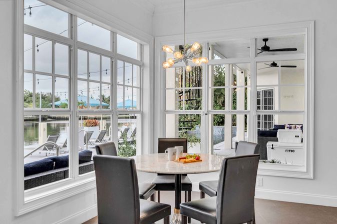 Airy waterfront dining nook with round marble table and four black chairs, large paned windows overlooking a dock, pool and string-lit patio