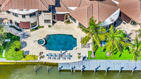Aerial view of a waterfront Mediterranean-style villa with terracotta roof, curved swimming pool, palm trees, sun loungers and a private dock on a canal