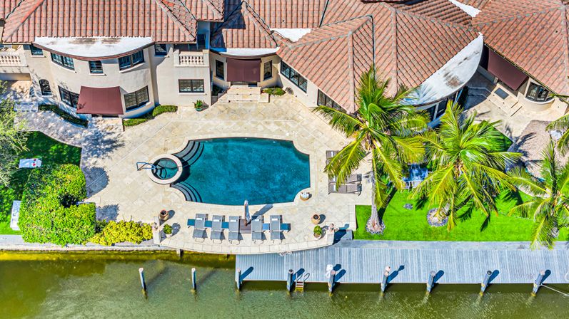 Aerial view of a waterfront Mediterranean-style villa with terracotta roof, curved swimming pool, palm trees, sun loungers and a private dock on a canal