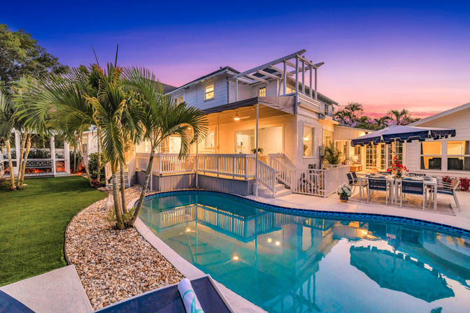 Tropical backyard at twilight with a lit two-story home, wraparound deck, palm trees, pebble-lined landscaping, outdoor dining under a navy umbrella, and a curved swimming pool reflecting the sunset.
