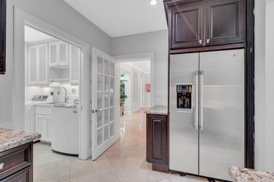 Bright modern kitchen with stainless steel side-by-side refrigerator, dark wood cabinets, granite countertops and tile floor; glass French door opens to a white laundry room with washer, sink and louvered cabinets.