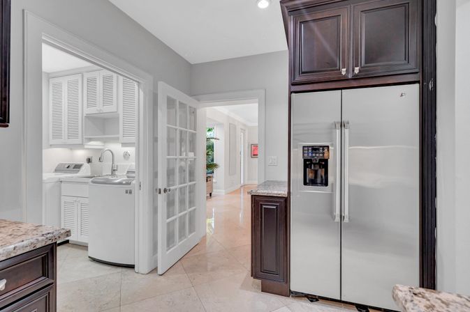 Bright modern kitchen with stainless steel side-by-side refrigerator, dark wood cabinets, granite countertops and tile floor; glass French door opens to a white laundry room with washer, sink and louvered cabinets.