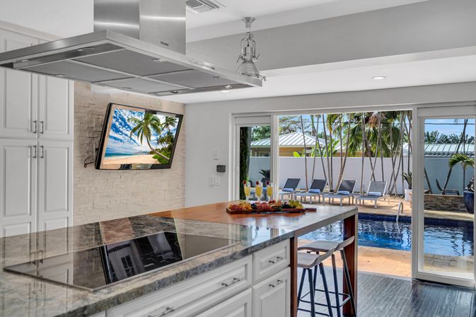 Bright modern kitchen with marble-topped island, bar stools and stainless range hood opening through sliding glass doors to a tropical poolside patio with lounge chairs and palm trees; wall-mounted TV shows a beach scene.
