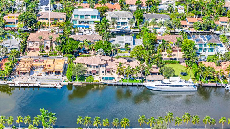 Aerial view of a tropical waterfront neighborhood with luxury homes, palm trees, private docks and a large white yacht moored on a calm canal, backyard pools and ongoing construction visible.