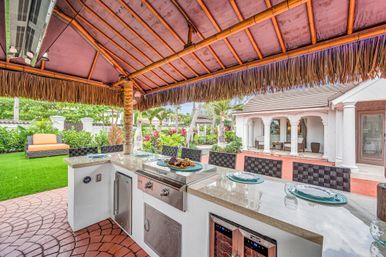 Tropical backyard outdoor kitchen under a tiki-style thatched palapa with a built-in stainless steel grill, marble countertop bar with place settings, wine cooler and woven bar stools, lush landscaping and patio seating by a stucco villa.