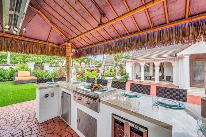Tropical backyard outdoor kitchen under a tiki-style thatched palapa with a built-in stainless steel grill, marble countertop bar with place settings, wine cooler and woven bar stools, lush landscaping and patio seating by a stucco villa.