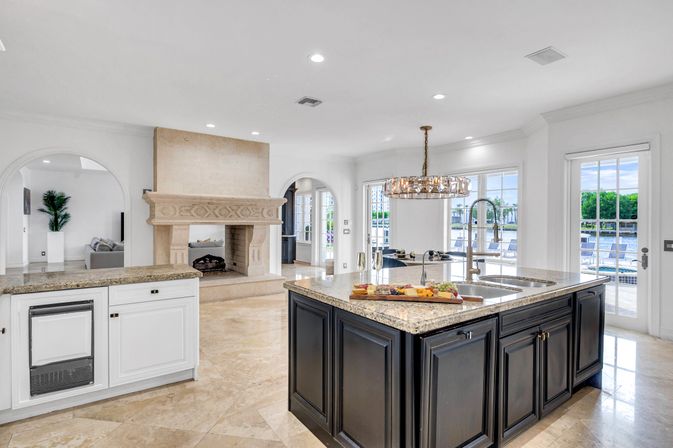 Bright waterfront luxury open-plan kitchen with large granite-topped island and sink, dark cabinetry, crystal chandelier, ornate stone fireplace, arched doorways and French doors opening to pool and dock views.