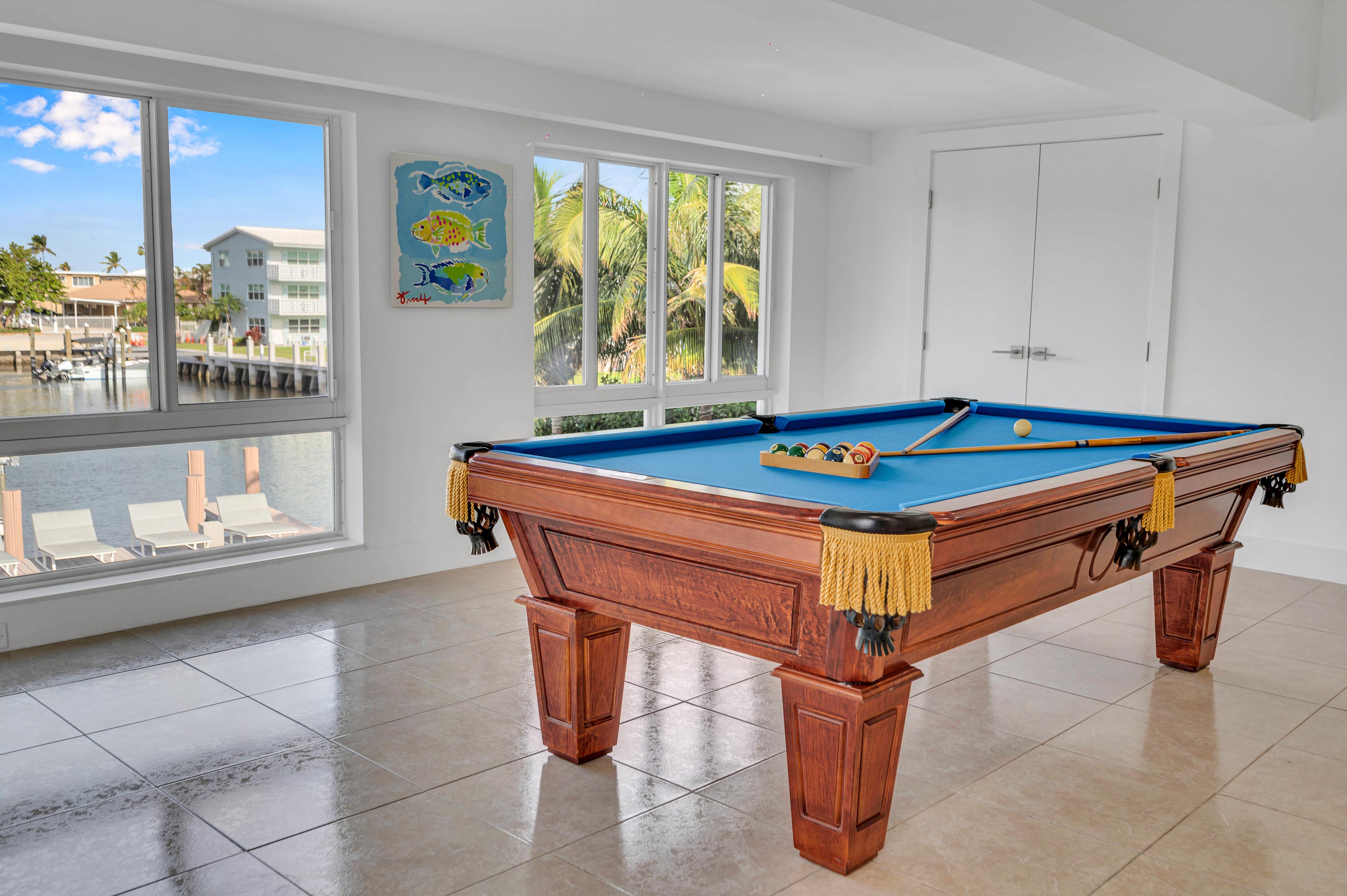 Sunlit coastal game room with a wooden pool table topped in blue felt and gold tassels, cues and racked balls on a tiled floor, large windows framing palm trees, boat docks and waterfront homes.