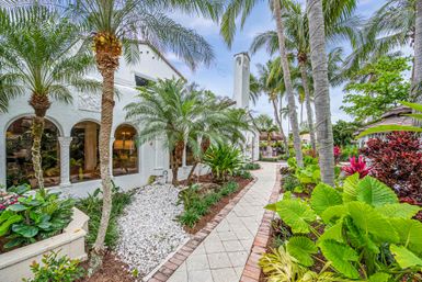 Paved palm-lined walkway beside a white stucco Mediterranean-style home with arched windows, lush tropical landscaping, and vibrant foliage