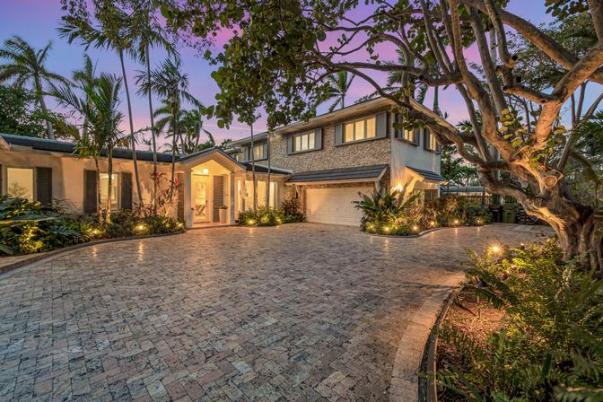 Twilight view of a two-story luxury home with stone façade and attached garage, warm entry lights, palm-lined tropical landscaping and a wide paver driveway beneath a pink-purple sunset.