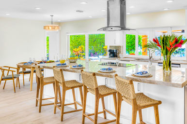 Bright, open-plan modern kitchen with marble island, stainless steel range hood, four woven rattan bar stools, table place settings, and sliding glass doors revealing a lush tropical backyard and pool.