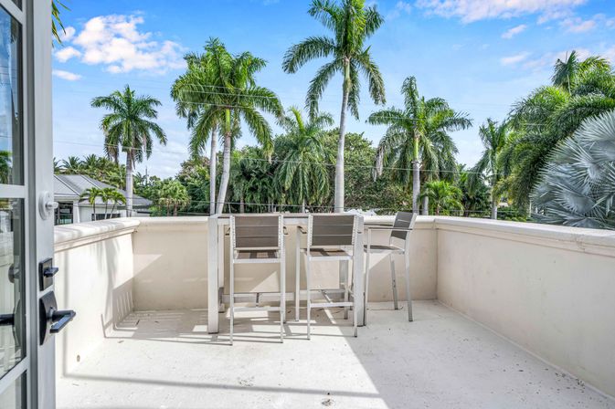 Sunny balcony with bar-height table and three stools overlooking tall palm trees and bright blue tropical sky, outdoor seating and residential view.