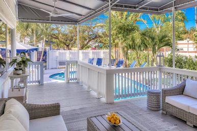 Bright tropical backyard patio with wicker sofas and ceiling fan, white railing and coffee table, overlooking a blue swimming pool, sun loungers and palm trees under a sunny sky.