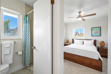 Sunny coastal-style bedroom with king bed and white linens, wooden frame, ceiling fan and wicker-lamp nightstands, seen through an open door to an en-suite bathroom with a teal patterned shower curtain, white towels and a window with palm trees.