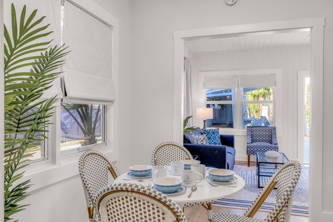 Cheerful sunlit coastal-style dining nook with a round white table set for four, woven bistro chairs and blue-and-white dishware; open view to a living room with navy sofa, patterned accent chair and tropical palms visible through the windows.