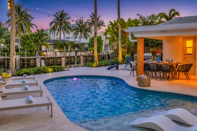 Tropical waterfront backyard with curved swimming pool, in-pool loungers and chaise lounges, string lights and palm trees at sunset beside a canal, covered dining patio and cozy outdoor seating.