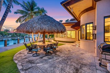Luxury tropical waterfront patio at sunset with a thatched palapa over a round dining table and swivel chairs, palm trees and dock along a calm canal, stone terrace, outdoor grill and warm-lit Mediterranean-style home.