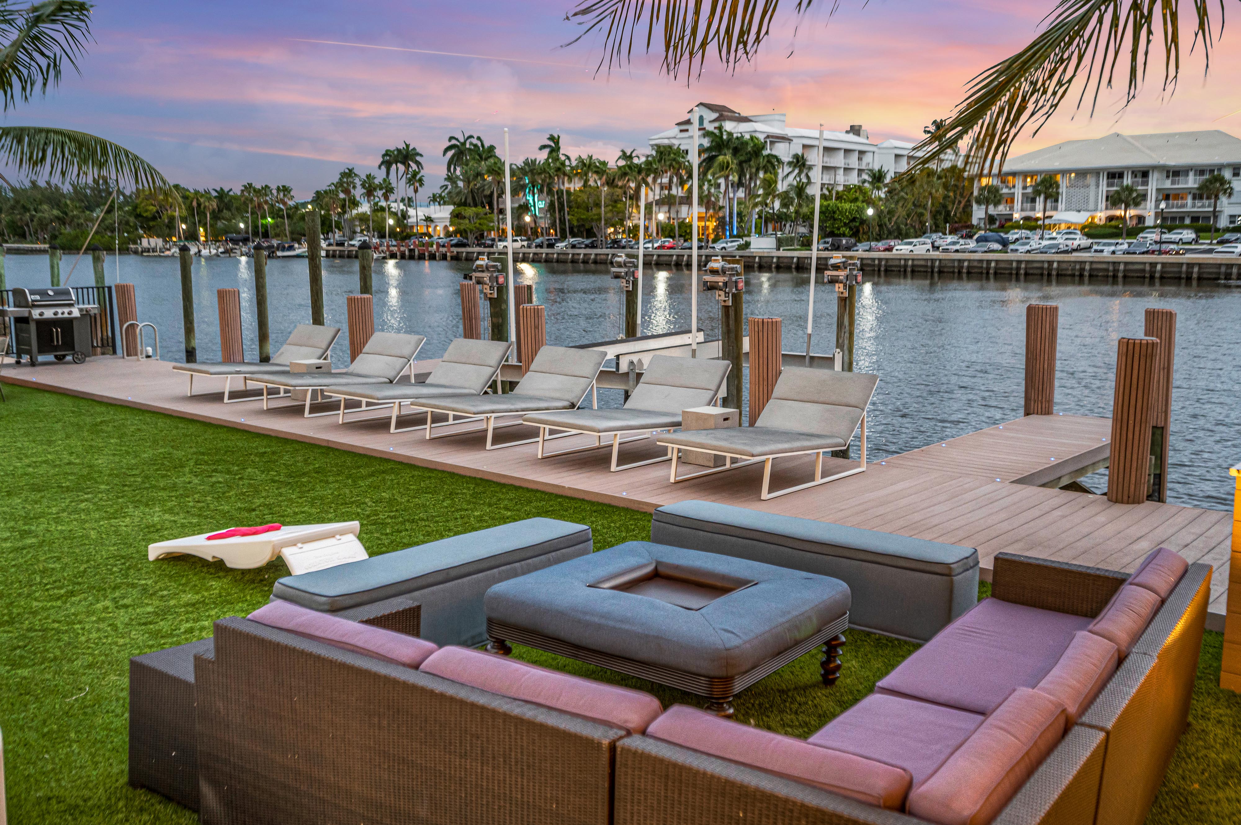 Sunset waterfront dock with palm trees, wooden pier and a row of sun loungers; cozy outdoor sectional on artificial turf overlooking a tropical marina and waterfront condos.