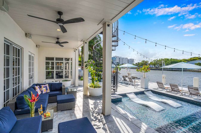 Relaxing sunny waterfront patio with navy sectional seating, mosaic plunge pool with in-pool loungers, spiral staircase, string lights and dock chairs along a calm canal