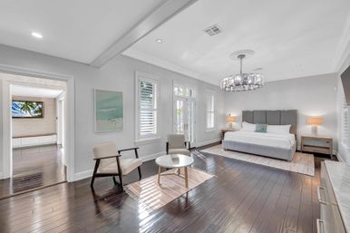 Spacious bright master bedroom with king bed and gray upholstered headboard, crystal chandelier, dark hardwood floors, seating area with two mid-century chairs and round coffee table, French doors and white plantation shutters.