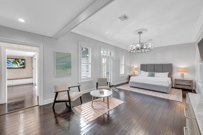 Spacious bright master bedroom with king bed and gray upholstered headboard, crystal chandelier, dark hardwood floors, seating area with two mid-century chairs and round coffee table, French doors and white plantation shutters.