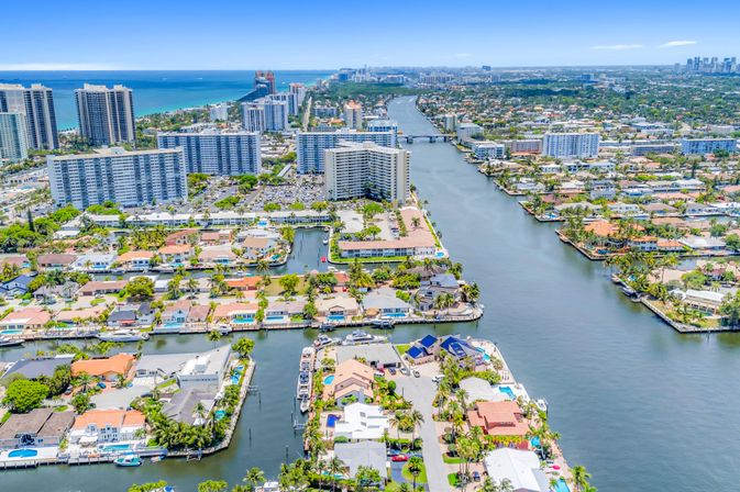Aerial view of a sunny Florida waterfront neighborhood with canals, palm-lined docks and boats, low-rise homes and oceanfront high-rise condos on the coastal skyline.