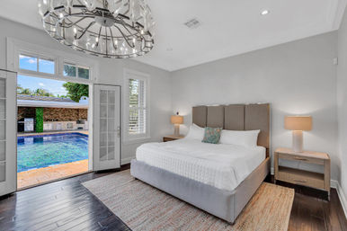 Modern master bedroom with king bed, chandelier and dark hardwood floors, French doors opening to a backyard pool and outdoor kitchen