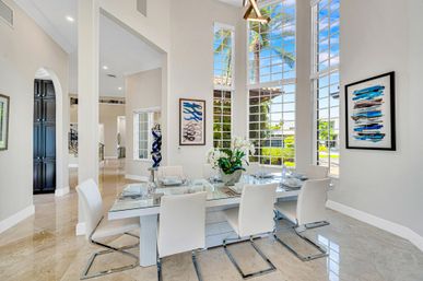 Sunny tropical dining room in a modern coastal home — glass-top table set for six with white leather chairs, tall grid windows framing palm trees and blue sky, blue abstract art and polished marble floors.