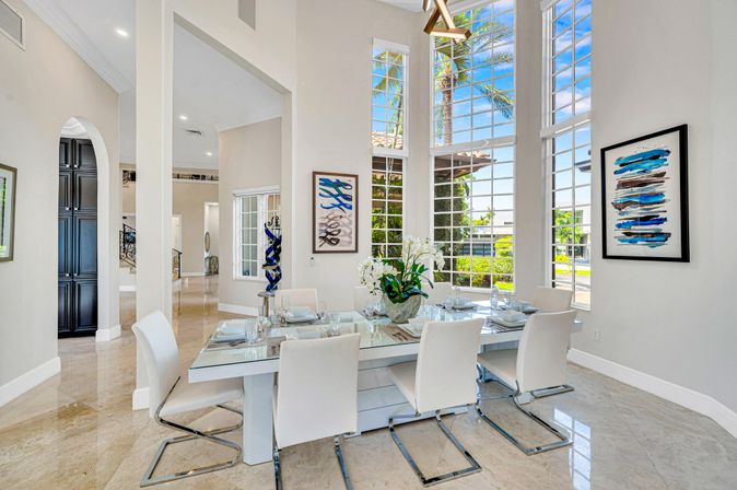 Sunny tropical dining room in a modern coastal home — glass-top table set for six with white leather chairs, tall grid windows framing palm trees and blue sky, blue abstract art and polished marble floors.