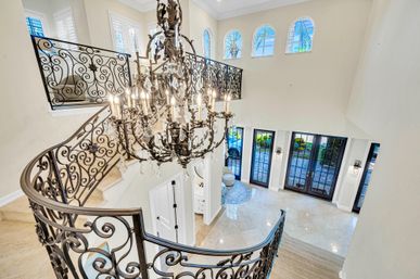 Sunlit luxury foyer with a sweeping curved marble staircase, ornate wrought-iron railing, sparkling crystal chandelier, arched clerestory windows, and double glass entry doors.