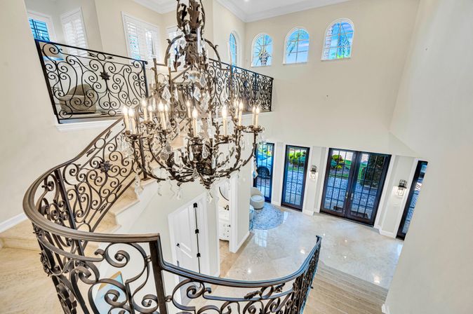 Sunlit luxury foyer with a sweeping curved marble staircase, ornate wrought-iron railing, sparkling crystal chandelier, arched clerestory windows, and double glass entry doors.