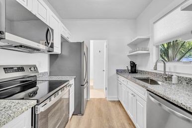 Bright modern white galley kitchen with stainless steel appliances, granite countertops, undermount sink, hardwood floor and a window framed by palm fronds.