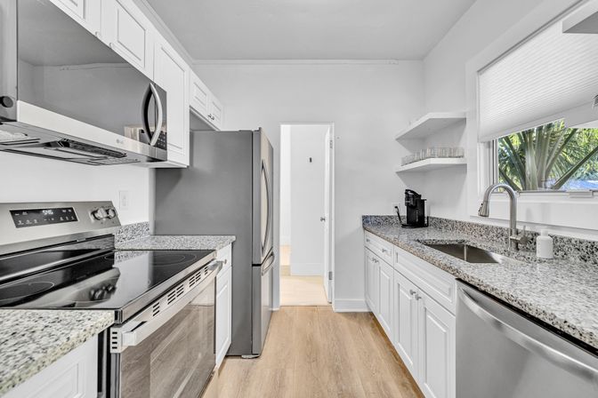 Bright modern white galley kitchen with stainless steel appliances, granite countertops, undermount sink, hardwood floor and a window framed by palm fronds.