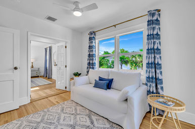 Bright coastal living room with white slipcovered sofa and navy throw pillows, blue palm-print curtains framing a window with tropical greenery and water view, rattan side table with magazine, patterned area rug, hardwood floors, ceiling fan, and sunlit French-door entry to adjoining room.