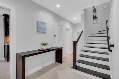 Modern home foyer with curved dark-wood staircase, slim console table holding a small potted plant, light tile floor, white walls, coastal wall art and recessed lighting.