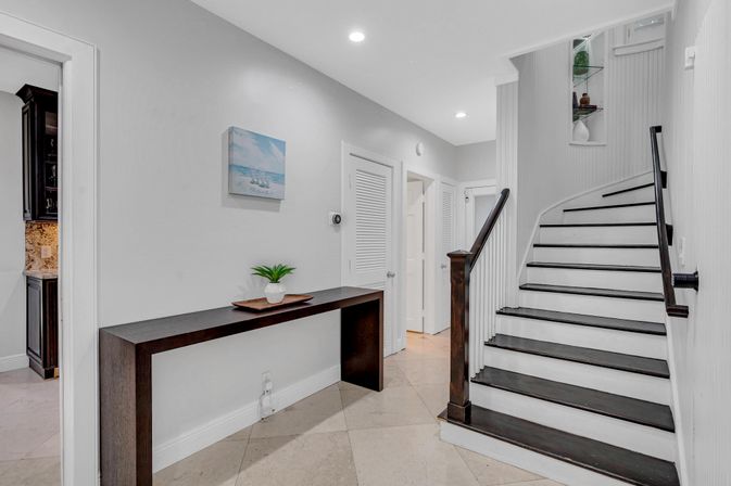 Modern home foyer with curved dark-wood staircase, slim console table holding a small potted plant, light tile floor, white walls, coastal wall art and recessed lighting.