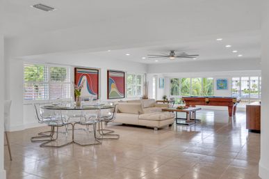 Bright open-plan living and dining room in a modern waterfront condo with clear acrylic chairs, beige sectional sofa, pool table, and large windows framing palm trees.