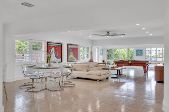 Bright open-plan living and dining room in a modern waterfront condo with clear acrylic chairs, beige sectional sofa, pool table, and large windows framing palm trees.