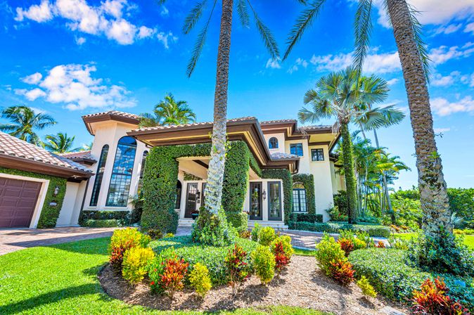 Mediterranean-style luxury coastal home with terracotta roof, tall palm trees, manicured tropical landscaping and bright blue sky