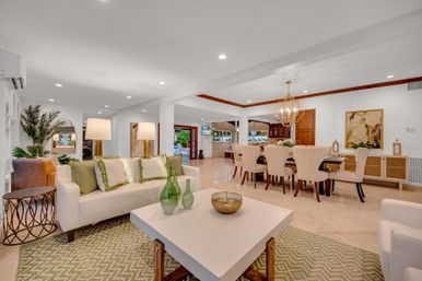 Sunlit open-plan living and dining room with white sofa and green accent pillows, patterned rug, white coffee table with green glass vases, chic gold chandelier over a beige dining table, indoor plants and sliding doors to a patio.