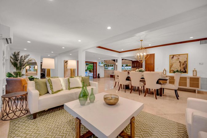 Sunlit open-plan living and dining room with white sofa and green accent pillows, patterned rug, white coffee table with green glass vases, chic gold chandelier over a beige dining table, indoor plants and sliding doors to a patio.