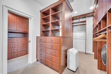 Spacious residential walk-in closet with custom dark wood built-in shelving and drawer island, hanging rods with wooden hangers, gray tile floor, recessed lighting, and a portable dehumidifier.