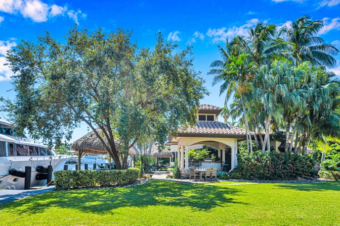 Sunny tropical waterfront gazebo and outdoor dining area beside a marina with docked yachts, palm trees, lush green lawn and bright blue sky.