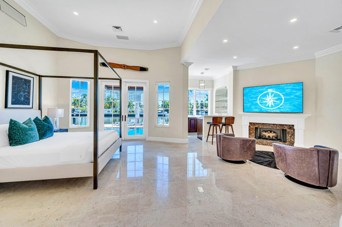 Sunlit luxury waterfront master suite with four-poster bed and teal pillows, marble floors, French doors opening to a private pool and marina framed by palm trees, cozy seating area with fireplace and wall TV, plus a small bar with stools.