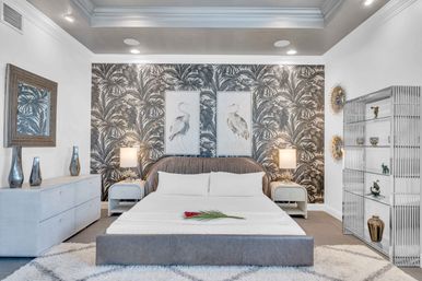 Modern tropical-inspired master bedroom with grayscale palm-print accent wall, king bed with upholstered headboard and white linens, twin heron prints, bedside lamps, dresser and open display shelving.