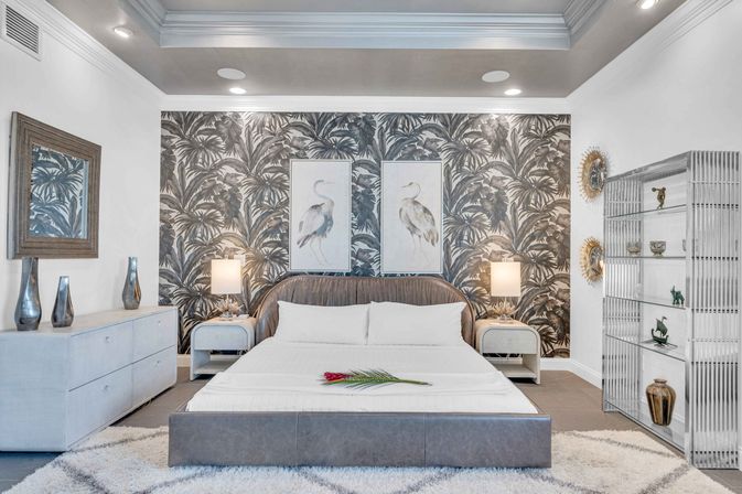 Modern tropical-inspired master bedroom with grayscale palm-print accent wall, king bed with upholstered headboard and white linens, twin heron prints, bedside lamps, dresser and open display shelving.