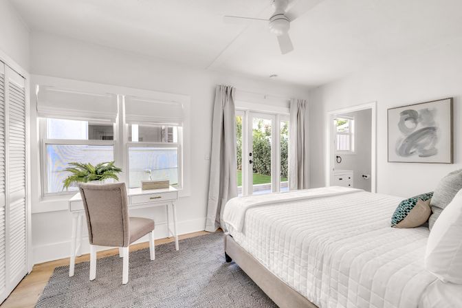 Airy sunlit modern bedroom with white quilted bed, patterned pillows, small white desk and chair by a window with a potted fern, French doors opening to a green backyard, neutral rug and abstract wall art.
