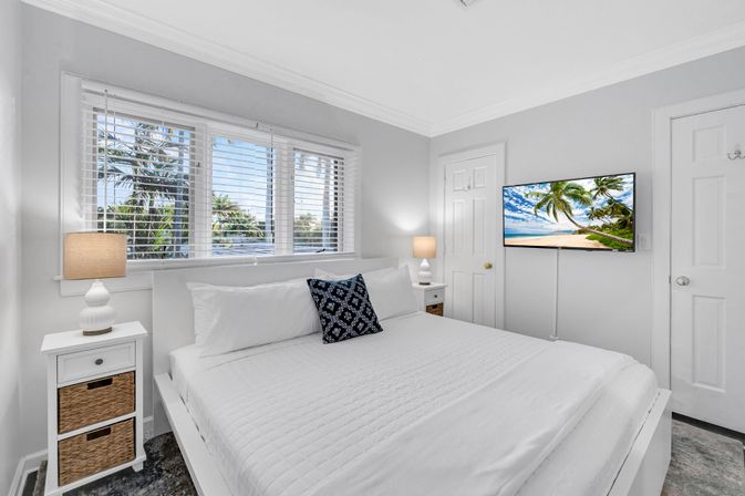 Bright coastal bedroom with white quilted king bed, navy patterned accent pillow, matching bedside tables and lamps, large window framing palm trees and ocean view, and wall-mounted TV showing a tropical beach scene.