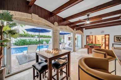 Sunlit indoor-outdoor game room with arched stone openings and exposed wood beams, featuring a chess table with stools, a pool table, bar seating, and sliding doors opening to a poolside patio with blue umbrellas and lounge chairs.