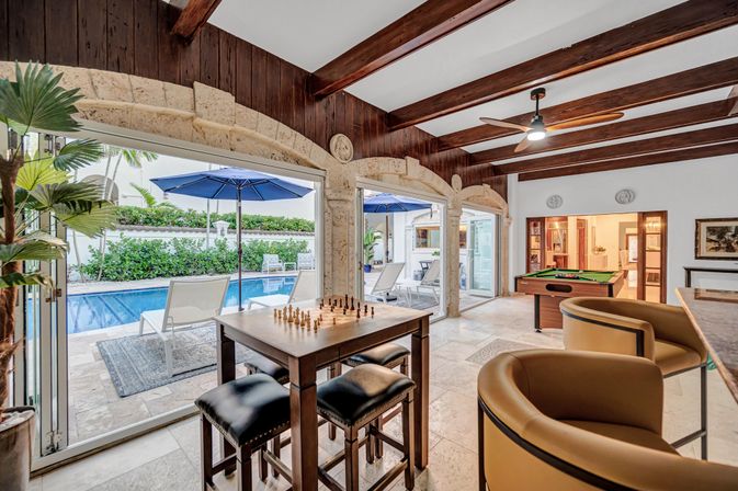 Sunlit indoor-outdoor game room with arched stone openings and exposed wood beams, featuring a chess table with stools, a pool table, bar seating, and sliding doors opening to a poolside patio with blue umbrellas and lounge chairs.